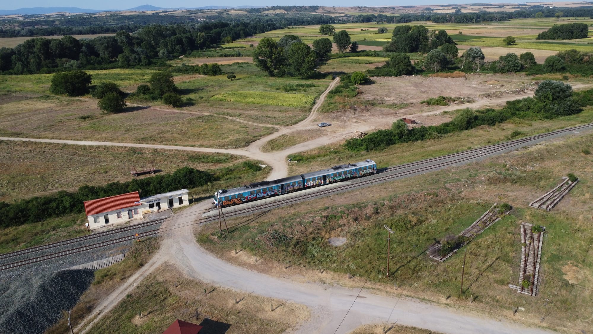 A DMU covered in graffiti stands at a tiny station. Odds and ends of track are to the sides of it, and fields beyond.